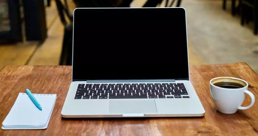 Ein aufgeklappter Laptop auf einem Tisch. Links daneben ein Notizblock mit Stift. Rechts vom Laptop eine volle Tasse Kaffee