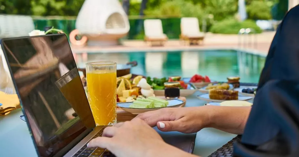 Eine Person tippt auf einem Laptop. Der Laptop steht auf einem Tisch mit Saft und Gurken. Im Hintergrund eine Poolanlage